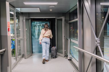 Riga, Letonya - 22 Şubat 2025 - bir kadın havaalanı jet köprüsünden bavulunu çekerek uçağına doğru ilerliyor. Modern terminal ayarı ve gece ışıkları bir seyahat atmosferi yaratır..