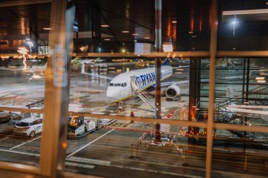 Riga, Letonya - 22 Şubat 2025 - Bir Ryanair uçağı gece havaalanı kapısına parketmiş, cam pencereden yansımalı olarak görülmüş. Yer araçları ve pist ışıkları yoğun bir atmosfer yaratır..