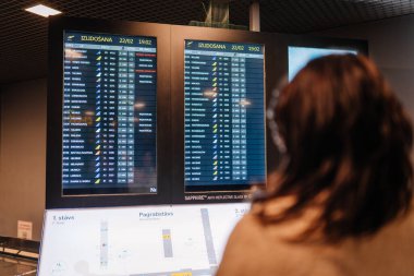 Riga, Letonya - 22 Şubat 2025 - Bir gezgin uçuş saatlerini, varış noktalarını ve geçit bilgilerini gösteren havalimanı kalkış panosunu kontrol eder. Sahne dijital ekranlarla aydınlatılıyor.