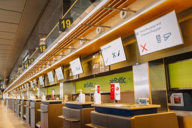 Riga, Letonya - 22 Şubat 2025 - AirBaltic markası, bagaj politikası ekranları ve numaralandırılmış tabelaları olan boş havaalanı kayıt gişeleri. Dezenfektanlar ve havayolu tabelası görünür..