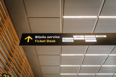 Riga, Letonya - 22 Şubat 2025 - Havaalanı bilet masası logolu, yolcuları yöneten. Letonya ve İngilizce genel tabelası, modern tavan ve arka planda ışıklandırma.