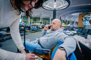 İstanbul, Türkiye - 23 Şubat 2025 - Gri kapüşonlu kel bir adam havaalanı bekleme salonunda uzanırken telefonla konuşuyor. Yakınlarda bir kadın bavulun üzerine eğiliyor..
