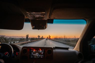 Antalya, Türkiye - 22 Şubat 2025 - Arka planda palmiye ağaçları ve dağlarla bir otoyolda gün batımına doğru giden bir arabanın içinden manzaralı bir yol manzarası.