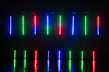 Koyu bir alanda kırmızı, mavi ve yeşil parlayan dikey neon ışıklar parlak zemini yansıtıyor, fütürist ve canlı bir sahne yaratıyor..
