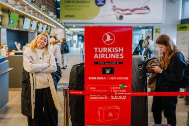 Riga, Letonya - 22 Şubat 2025 - Türk Hava Yolları kayıt alanındaki yolcular, havaalanındaki kırmızı bagaj politikası tabelasının yanında duruyor, arka planda yoğun bir terminal bulunuyor..