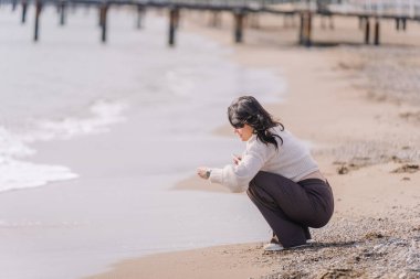 Suya yakın kumsalda çömelmiş bir kadın, elinde bir telefon, kumsala dokunuyor, deniz kenarında huzurlu bir anın tadını çıkarıyor..