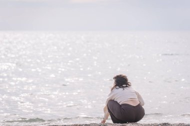 Bir kadın çakıl taşı plajında çömelmiş, yumuşak, dağınık bir ışığın altında parıldayan denize bakıyor. Huzurlu ve huzurlu sahil manzarası.