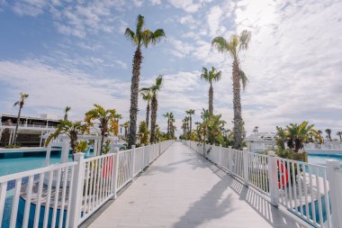 Palmiye ağaçlarıyla çevrili beyaz bir köprü, parlak mavi bir gökyüzünün altındaki lüks bir tatil köyünde, sakin ve tropikal bir atmosfer yaratıyor..