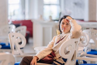 Rahat bir süveter giyen bir kadın şık bir kafede zarif beyaz bir sandalyede oturuyor, düşünceli bir ifadeyle başını elinin üzerine yaslıyor..