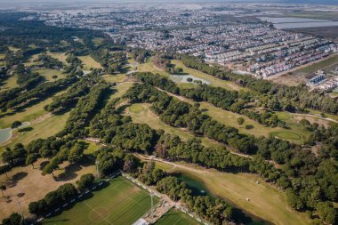 Bereketli yeşillikler, kum sığınakları, su tehlikeleri ve temiz bir gökyüzünün altında yakın bir şehir manzarası olan bir golf sahasının havadan görünüşü..