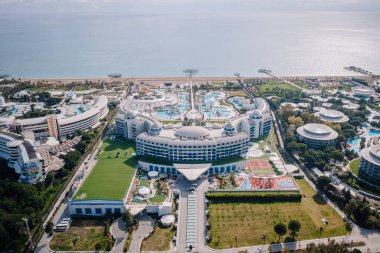 Serik, Türkiye - 28 Şubat 2025 - Havuzu, yemyeşil bahçeleri, spor tesisleri ve parlak bir gökyüzünün altındaki deniz manzaralı lüks bir sahil beldesi.