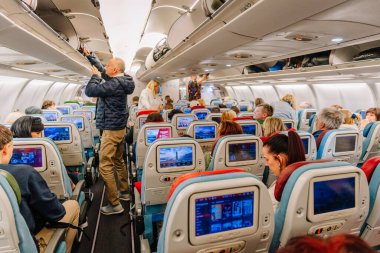Riga, Letonya - 22 Şubat 2025 - Bir uçak kabinindeki yolcular uçuş için hazırlanıyor, bazıları oturma odası eğlence ekranları kullanıyor ve diğerleri de üstlerinde bavul depoluyor..