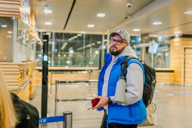 Riga, Letonya - 22 Şubat 2025 - Şapka takan ve gözlüklü bir erkek yolcu, elinde pasaport ve biniş kartıyla havaalanı kontrol noktasında bekliyor..