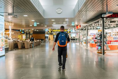 Riga, Letonya - 22 Şubat 2025 - Mavi ve turuncu sırt çantalı bir adam modern bir havaalanı terminalinde yürüyor, parlak ışık altında dükkanlardan ve oturma alanlarından geçiyor.