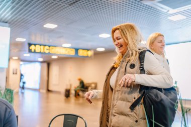 Riga, Letonya - 22 Şubat 2025 - Sırt çantalı iki gülümseyen kadın havaalanı terminaline vardıklarında yolculukları için mutlu ve heyecanlı görünüyorlardı. Arka planda bir çıkış işareti görünür.