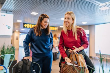 Riga, Letonya - 22 Şubat 2025 - Havaalanı terminalinde bavulları ve alışveriş çantalarıyla gülümseyen iki kadın. Uçuşlarını beklerken mutlu ve konuşarak meşgul görünüyorlar..