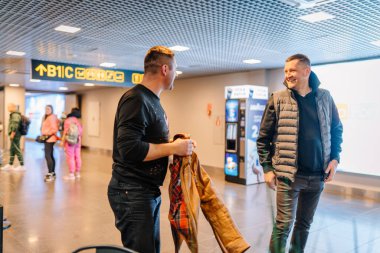 Riga, Letonya - 22 Şubat 2025 Havaalanı terminalinde gülümseyen ve konuşan iki adam. Biri ceketi tutarken, diğerleri arka planda yürür. Gitmeden önce dostça bir sohbet..