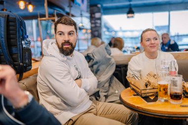 Riga, Letonya - 22 Şubat 2025 - Beyaz kapüşonlu sakallı bir adam bir kafe masasında kollarını kavuşturmuş otururken, gülümseyen bir kadın içkiden zevk alır. Bardaklar ve atıştırmalıklar masada..