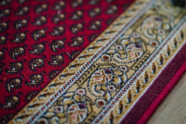 Geleneksel kırmızı ve altın desenli, karmaşık detaylarla döşenmiş bir halıya yakın çekim, ahşap bir zeminin dokusu ve işçiliği vurgulanıyor..