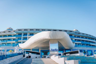 Serik, Türkiye - 1 Mart 2025 - Fütürist beyaz gölgelikli modern lüks otel girişi, mavi cam balkonları ve açık mavi gökyüzünün altında park edilmiş tur otobüsleri.