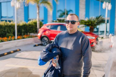 Serik, Türkiye - 1 Mart 2025 - Güneş gözlüğü ve koyu renk sweatshirt giyen bir adam, arkasında kırmızı bir araba, palmiye ağaçları ve modern bir bina bulunan bir su şişesi ve ceket tutuyor.