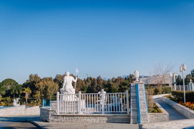 Serik, Türkiye - 1 Mart 2025 - Poseidon 'un üç dişli mızrak taşıyan beyaz heykeli yeşil, mavi gökyüzü ve yürüyüş yolu olan açık bir çitin arkasında oturuyor.