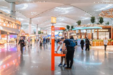 İstanbul, Türkiye - 01 Mart 2025 - Havaalanı terminalindeki yolcular turuncu dijital bilgi kioskuyla etkileşim halindeler. Modern terminalin lüks dükkanları ve parlak ışıkları var..