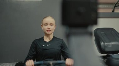 Valmiera, Letonya - 4 Şubat 2025 - Siyah spor giyim giymiş genç bir kadın spor salonunda spor salonuna odaklanmış kürek çekiyor. Arka planda spor aletleri var.