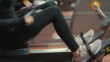 Valmiera, Letonya - 4 Şubat 2025 - Spor salonunda spor yaparken, siyah tayt ve beyaz spor ayakkabılar giyerek kürek çekme makinesine bağlanmış bir insan bacağı ve ayaklarının yakın çekimi.