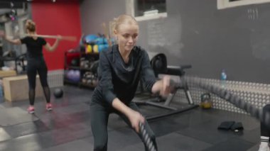 Valmiera, Letonya - 4 Şubat 2025 - odaklanmış bir kadın spor salonunda savaş halatlarıyla egzersiz yapar, dayanıklılık ve dayanıklılık sağlar. Arka planda çalışan başka biri daha var..