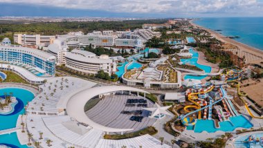 Serik, Türkiye - 28 Şubat 2025 - Havuz, su parkı slaytları, modern mimari, palmiye ağaçları ve manzaralı kıyı manzaralı lüks bir sahil beldesi manzarası.