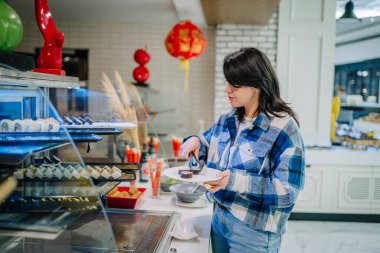 Belek, Türkiye - 22 Şubat 2025 - Mavi ekose gömlekli bir kadın modern bir restoranda bir açık büfeden suşi seçiyor. Dekorasyonda Asya 'dan esinlenilmiş bir dekor ve bir gıda sergisi yer alıyor.