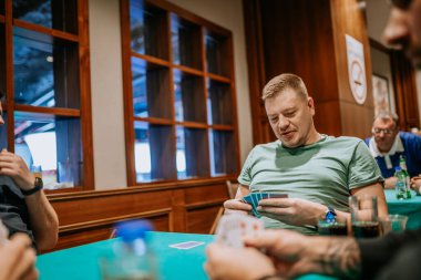 Belek, Türkiye - 22 Şubat 2025 - Yeşil tişörtlü bir adam sosyal ortamda kart oynuyor, elini tutarken gülümsüyor. Masada diğer oyuncular ve içkiler görünür..