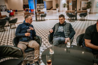 Belek, Türkiye - 22 Şubat 2025 - İki erkek şık bir salonda oturmuş, içki içerken sohbet ediyorlar, rahat bir atmosferi ve modern iç tasarımı var..