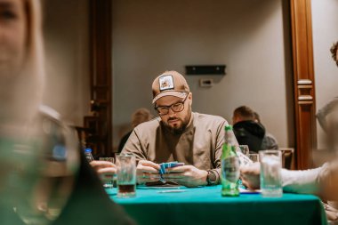 Belek, Türkiye - 22 Şubat 2025 - Şapkalı ve gözlüklü sakallı bir adam loş ışıklı bir odada yeşil bir masada kağıt oynuyor, elinde kağıt oynarken eline konsantre oluyor..