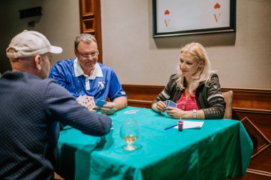 Belek, Türkiye - 22 Şubat 2025 - Üç kişi yeşil bir bezle kaplı bir masada kağıt oynuyor. Masada içkiler ve notlarla odaklanmış durumdalar..