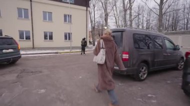 Valmiera, Letonya - 4 Şubat 2025 - Kürklü bir kadın karlı bir kaldırımda yürür, bir el çantası taşır. Arka planda ağaçlar, bir bina ve kışlık giysili başka bir yaya var..