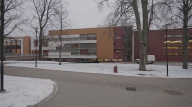 Valmiera, Letonya - 4 Şubat 2025 - Karlı bir kış gününde geniş cam pencereli ve kahverengi cepheli modern bir okul binası. Ön planda çıplak ağaçlar ve sokak lambaları duruyor..