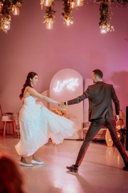 Koknese, Letonya - 26 Eylül 2023 - Bir gelin ve damat düğün törenlerinde neşeli bir şekilde dans ediyor, sıcak ışık ve çiçeklerle dolu zarif bir mekanda el ele tutuşuyorlar.