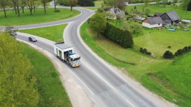 Valmiera, Letonya - 19 Mayıs 2024 - Eğri bir kırsal yolda modüler bir evi taşıyan bir kamyonun havadan görünüşü, bir çiftlik evi ve küçük bir arabayla yeşil bir araziyi geçiyor.