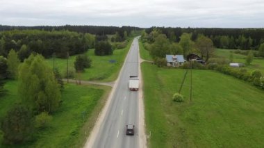 Valmiera, Letonya - 19 Mayıs 2024 - Yeşil tarlalar, ağaçlar ve geçen arabalarla çevrili kırsal bir otoyolda modüler ev taşıyan bir kamyonun havadan görünüşü.