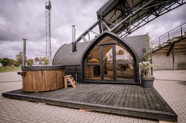 Valmiera, Letonya - 10 Mayıs 2024 - Güvertesinde ahşap jakuzisi olan modern siyah kemerli bir kulübe. Cam kapılar ve saksı bitkileriyle şık bir dinlenme yeri..
