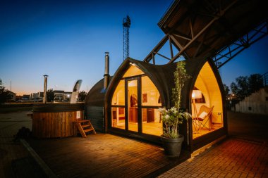 Valmiera, Letonya - 10 Mayıs 2024 - Alacakaranlıkta sıcak iç aydınlatma ile modern kemerli küçük bir ev, ahşap bir güverte, sıcak bir jakuzi ve rahatlama için rahat bir açık hava düzeneği.