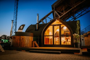 Valmiera, Letonya - 10 Mayıs 2024 - Alacakaranlıkta sıcak ışıklandırması olan, ahşap jakuzili ve şık dış dekorlu, rahat bir atmosfer yaratan modern bir ahşap kulübe.