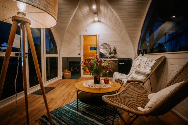 Sıcak ahşap kabin içinde hasır sandalyeler, kırsal bir sehpa, bir şömine, ve sıcak aydınlatma, rahatlatıcı ve davetkar bir ortam yaratıyor..