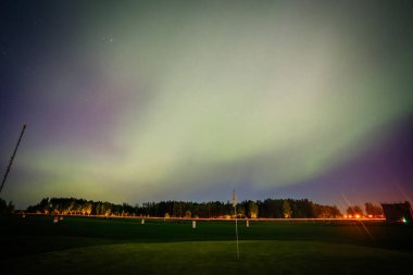 The northern lights glow over a golf course at night, with a green fairway, flag, and distant city lights creating a serene atmosphere. clipart