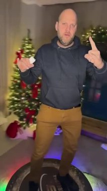 Valmiera, Letonya - 15 Aralık 2023 - Kapüşonlu kel bir adam 360 'lık bir video kabin platformunda, Noel süsleri ve festival ışıklarıyla çevrili bir şekilde taş el hareketleriyle poz veriyor..