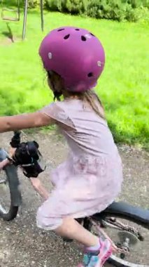 Valmiera, Letonya - 12 Ağustos 2023 - Pembe kask takan genç bir kız yeşil parktaki kirli bir yolda pembe bir bisiklete biniyor. Güneş ışığı ağaçların arasından süzülür, gölgeler bırakır..