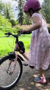 Valmiera, Letonya - 12 Ağustos 2023 - Pembe kask takan genç bir kız yeşil parktaki kirli bir yolda pembe bir bisiklete biniyor. Dantel bir elbise ve renkli ayakkabılar giyiyor..