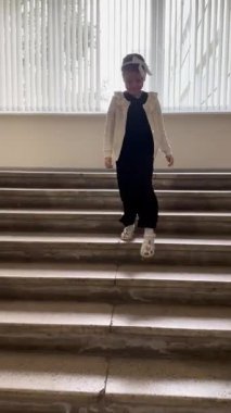 Şık giyinmiş genç bir kız, beyaz bir hırka, siyah bir tulum ve bir fiyonk takarak, parlak bir iç mekan ortamında merdivenlerden aşağı iniyor..